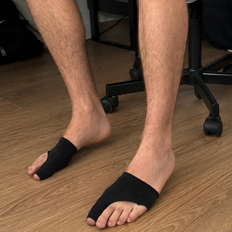 A person sitting on a black office chair in a home office setting, wearing black split-toe toe separators on their feet. The room features a wood floor, a desk with a laptop, and a black backpack in the background.